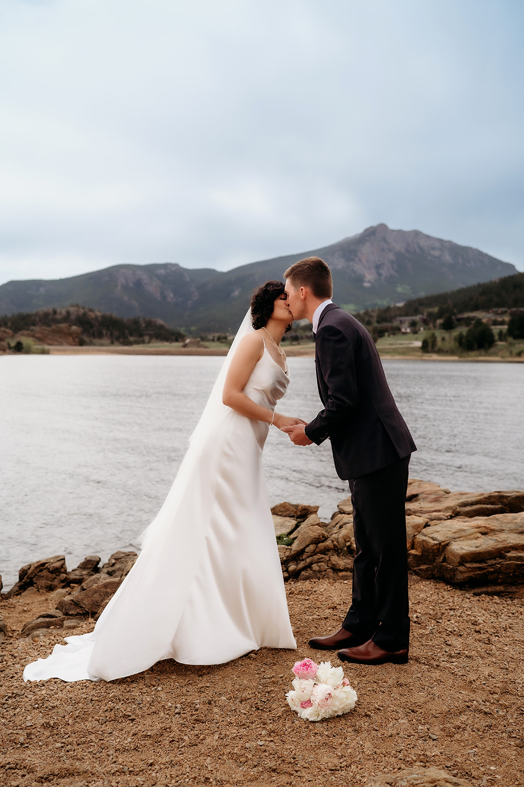 Just married couple shares a kiss lakeside with a stunning mountain backdrop, capturing the romance of Colorado elopement locations.