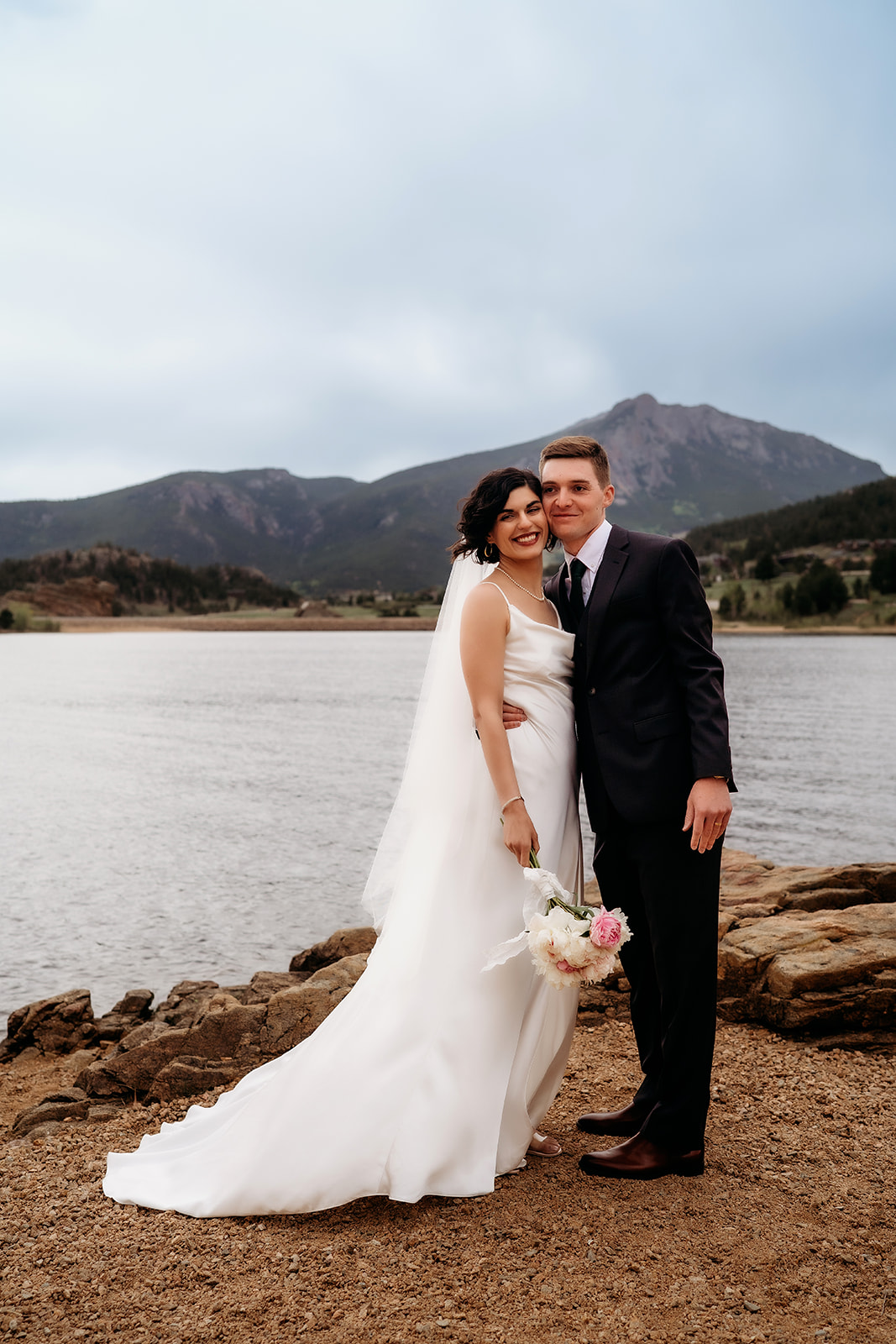 Bride and groom smile at the camera while standing lakeside, with mountain views framing this peaceful Colorado elopement location.