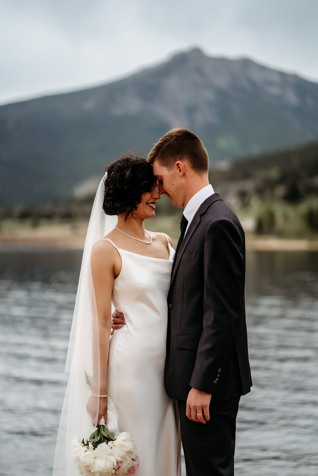 Bride and groom touch foreheads with soft smiles, bouquet in hand, as they savor a quiet moment by the lake after their ceremony.