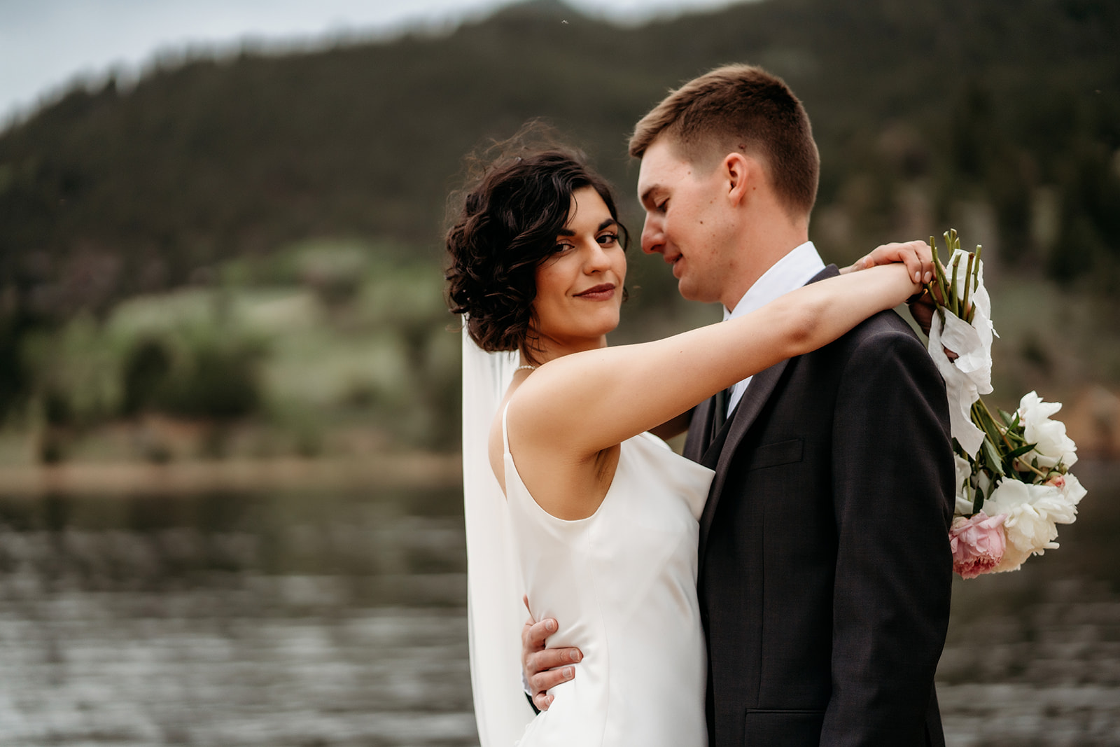Newlyweds embrace joyfully with soft florals in hand, surrounded by pine-covered mountains at a lakeside Colorado elopement location.