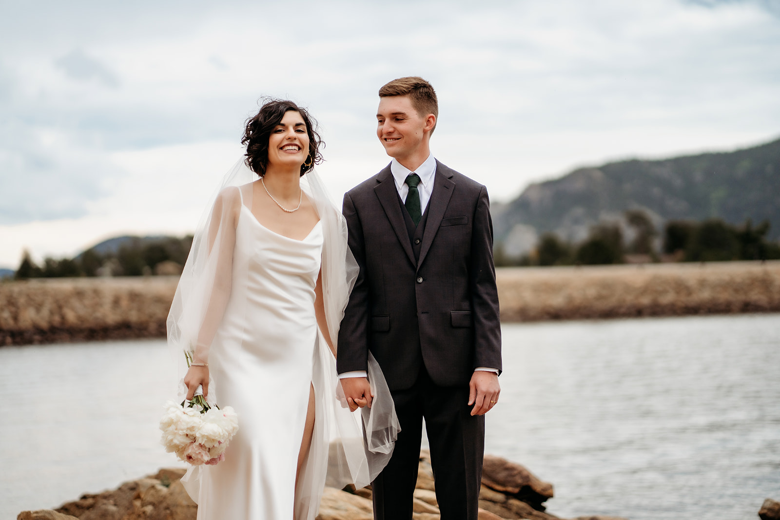 Bride and groom share a joyful walk beside the lake, her bouquet in hand and smiles wide under the cloudy Colorado sky.