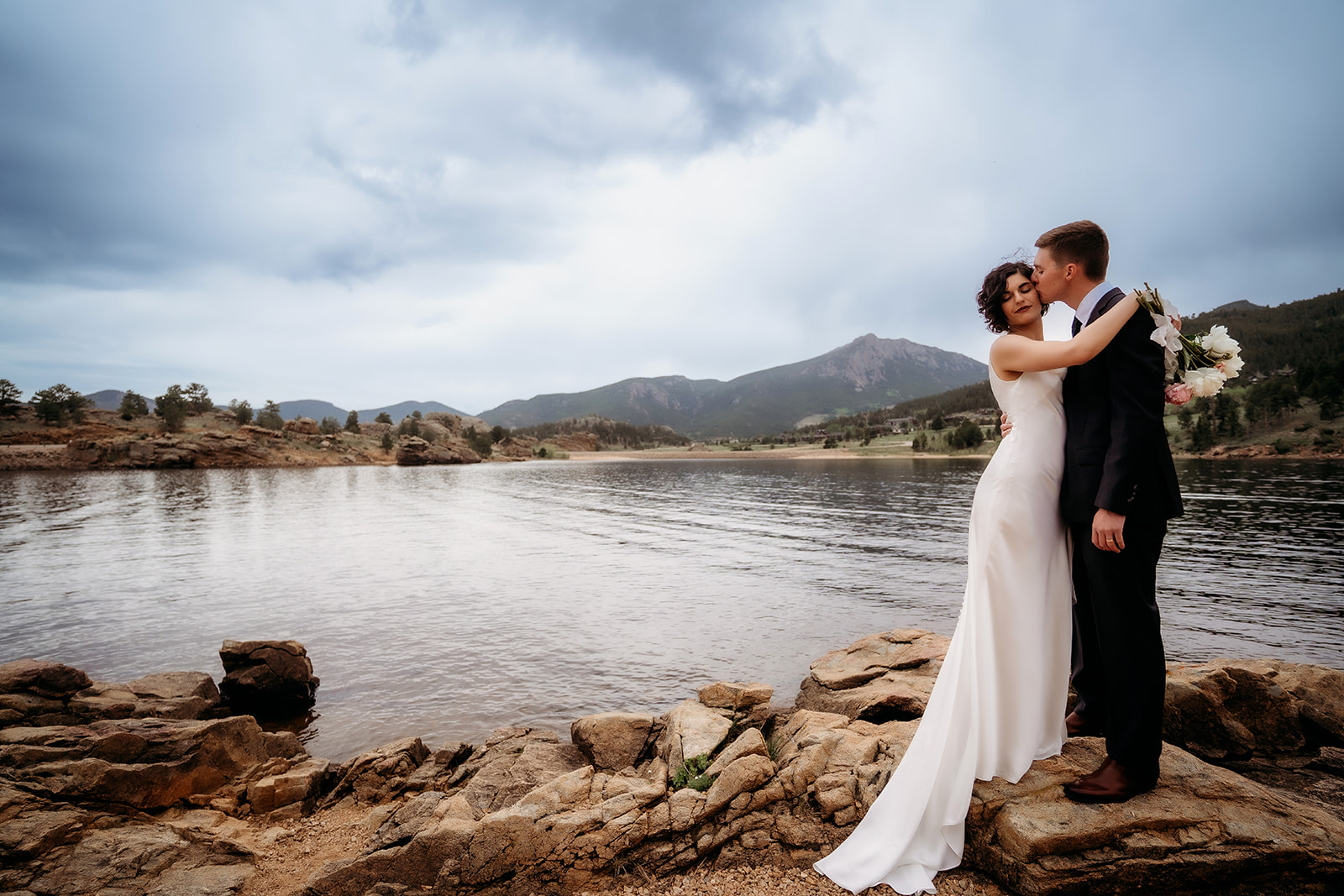 Bride wraps her arms around the groom as they stand on the rocks above the water, with Colorado mountains in the distance.