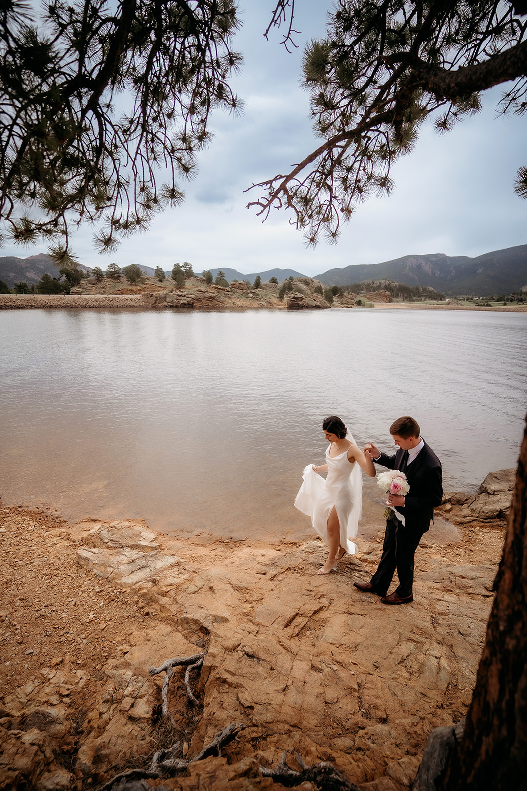 Groom helps bride down a rocky path by the water’s edge during their adventure elopement in Colorado.