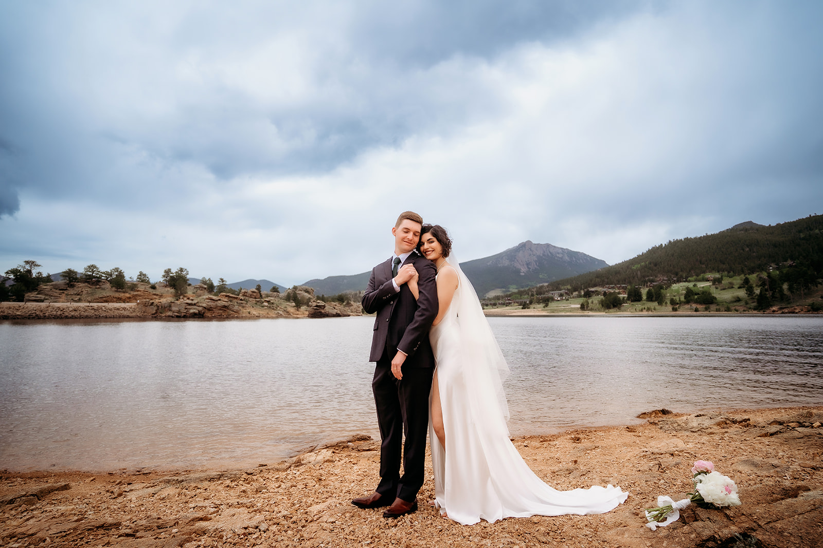 Bride leans on her groom with a soft smile as they pose along the rocky lakeshore after their Colorado elopement.