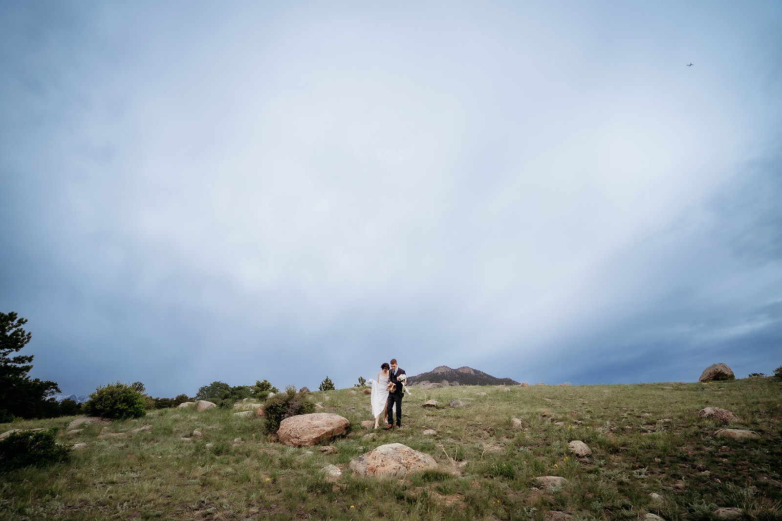 Wide, dramatic shot of the couple standing together in an open mountain field, dwarfed by sky and stone—a breathtaking Colorado elopement location.