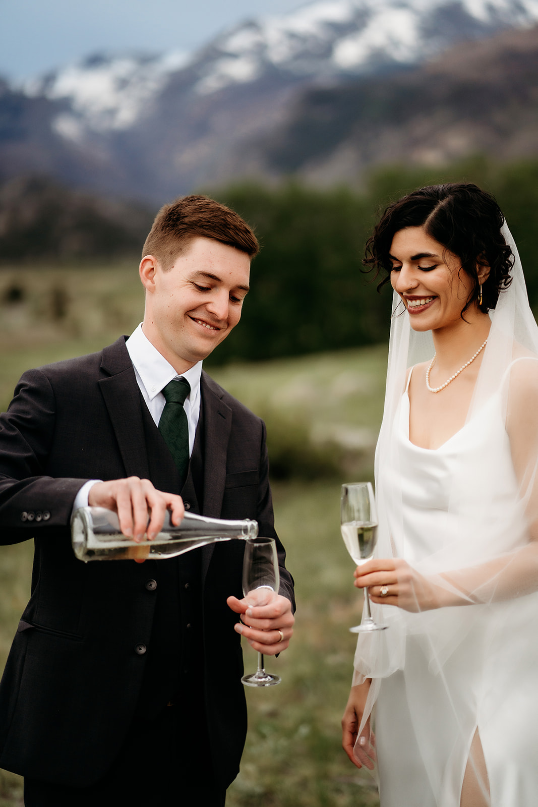 Groom pours champagne into a flute while the bride smiles, celebrating in a quiet meadow with the Rockies rising behind them.