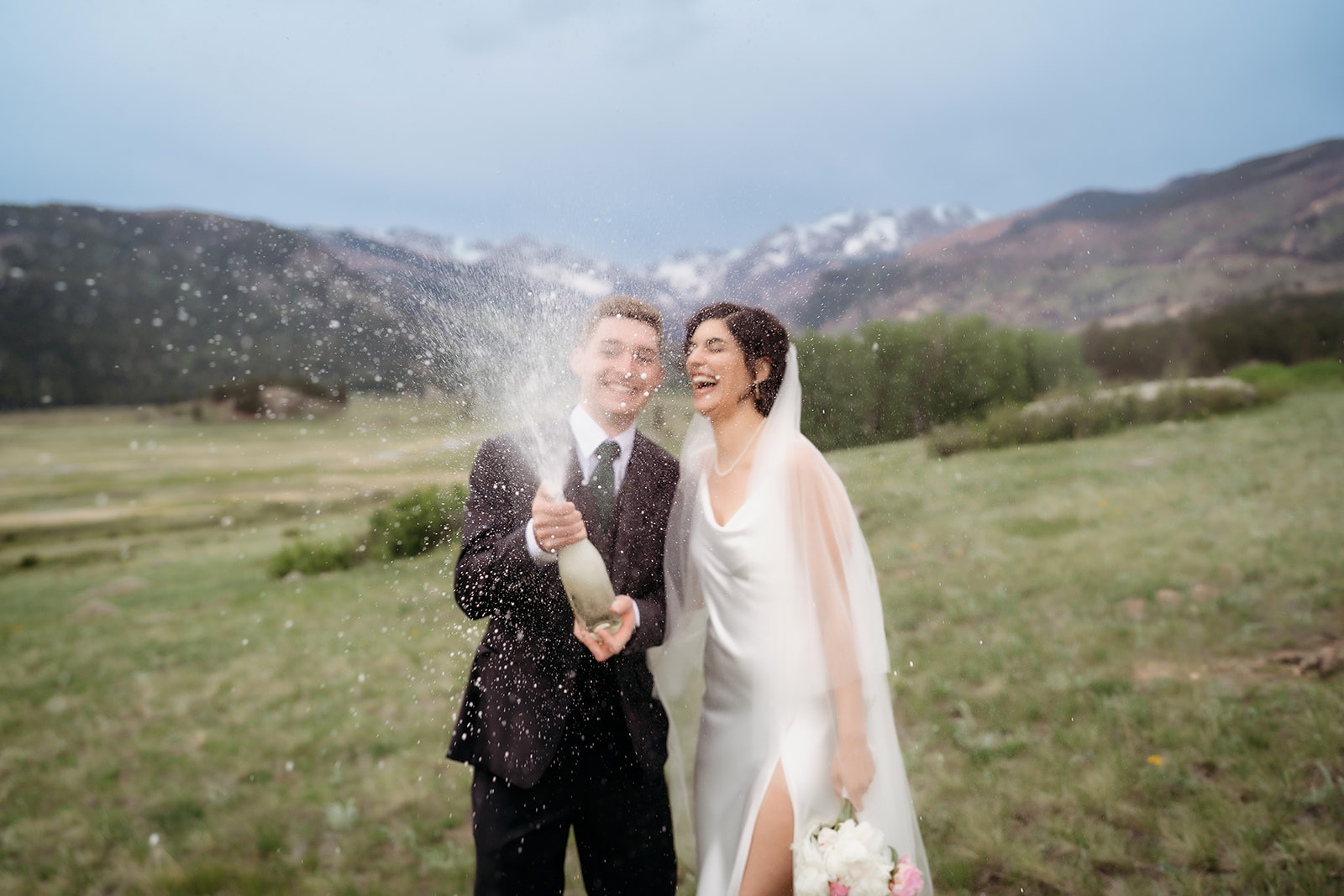 Bride and groom popping champagne and laughing in an open mountain meadow, one of the more joyful Colorado elopement locations.