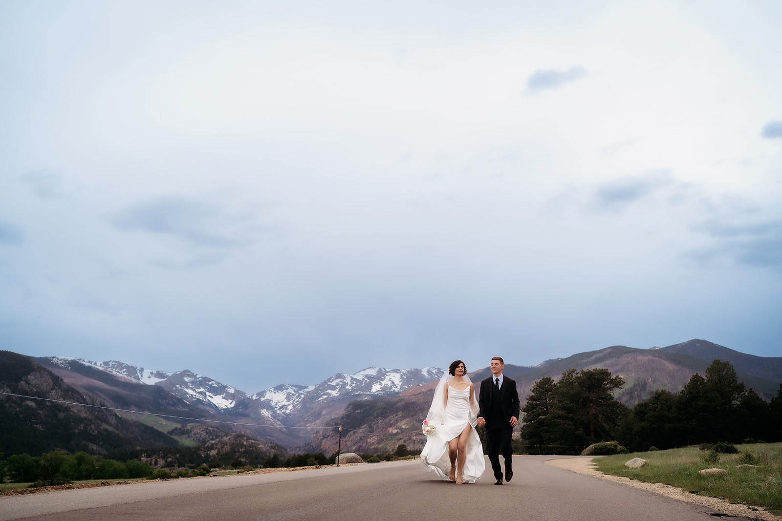 Wide-angle shot of the couple walking down the open road with snowcapped peaks behind them, highlighting the adventure of Colorado elopement locations.