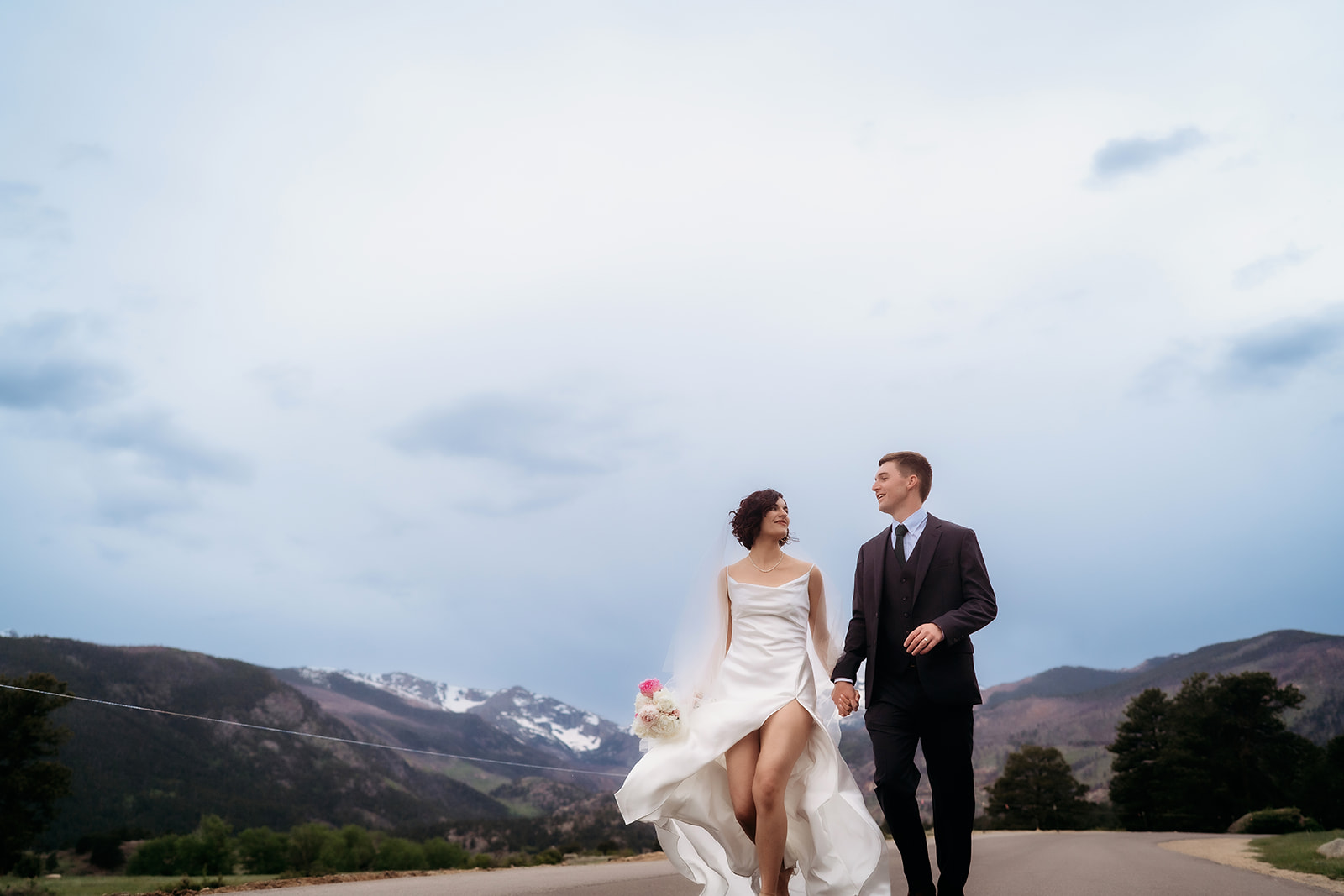 Bride and groom walking hand in hand along a mountain road, full of joy and wind-blown romance at one of the most scenic Colorado elopement locations.