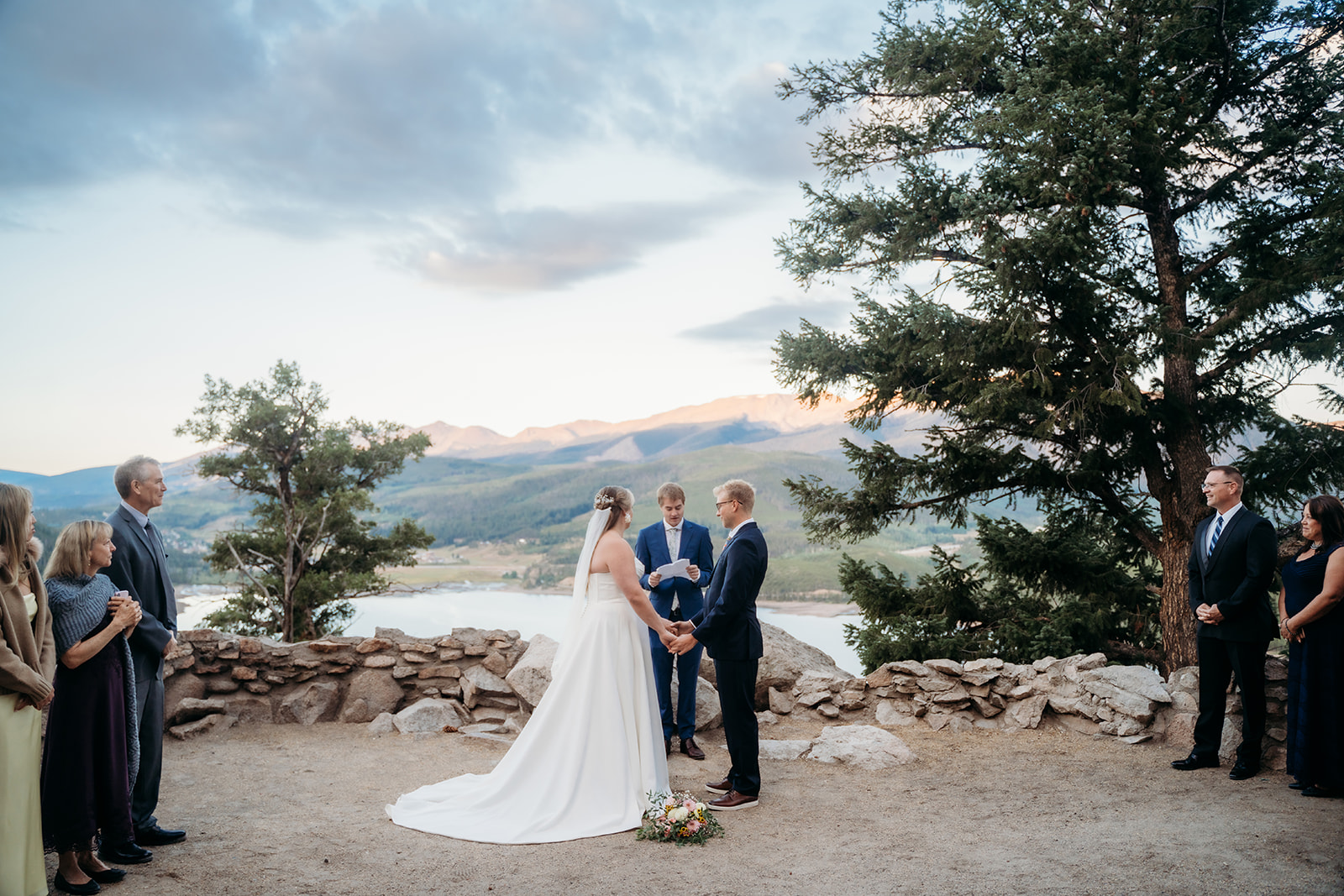 Wide shot of couple exchanging vows surrounded by family, mountains and lake in the background, by Denver elopement photographer.