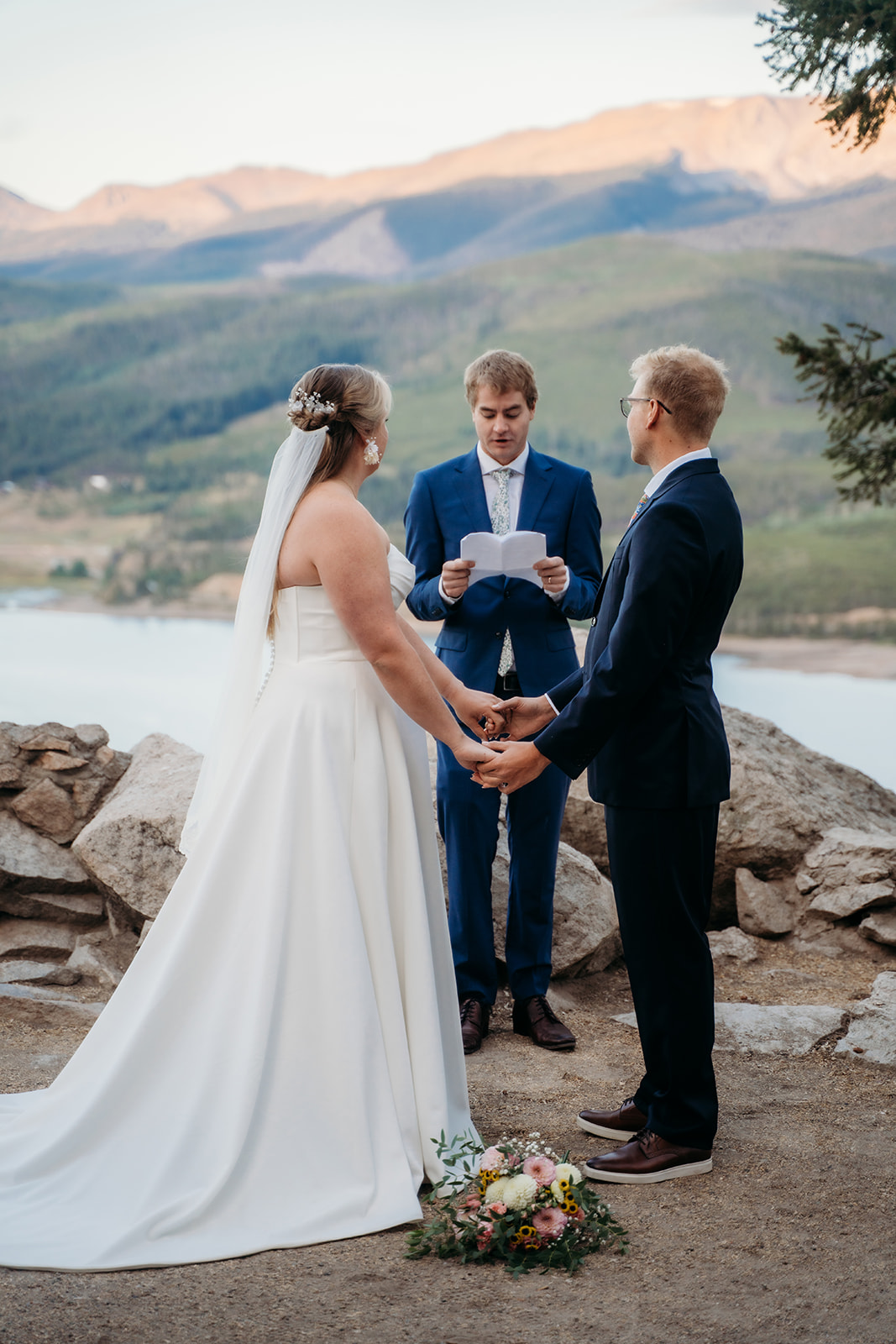 Bride and groom holding hands during their mountain ceremony with an officiant reading vows, captured by a Denver elopement photographer.