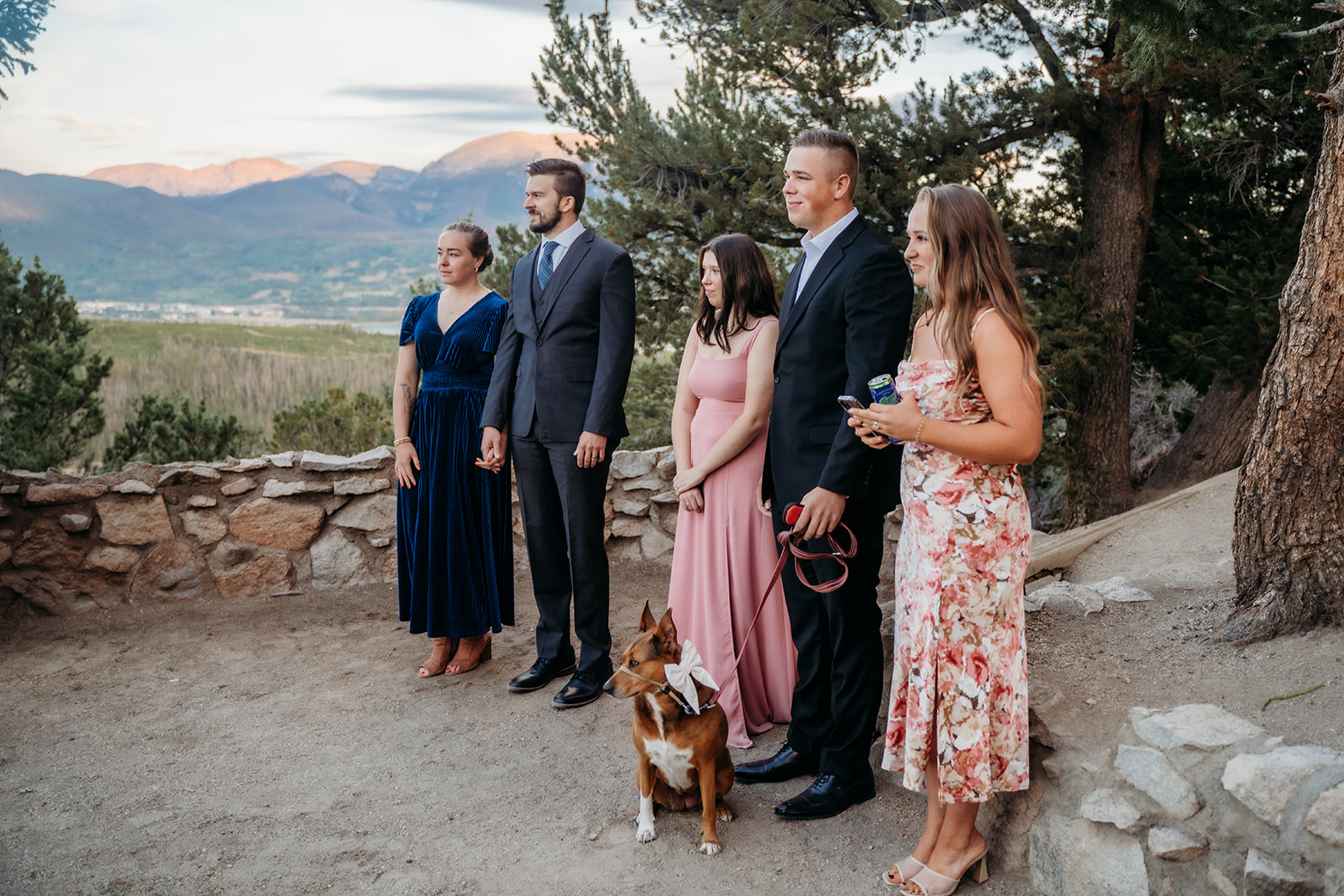 Wedding guests dressed in colorful attire standing with the couple’s dog, watching an outdoor mountain ceremony unfold, captured by a Denver elopement photographer.