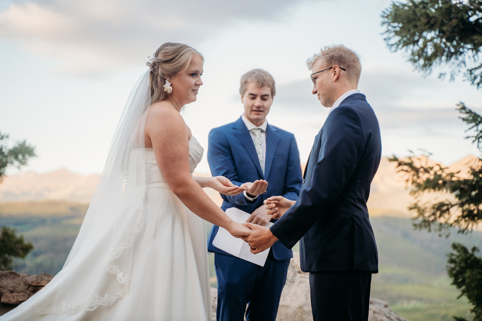 Bride and groom holding hands while exchanging vows under the trees with mountain views in the distance.