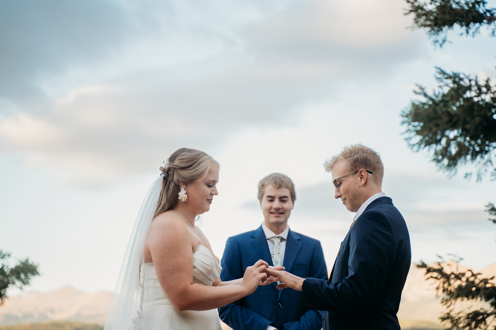 Groom placing ring on bride’s finger as officiant looks on during outdoor ceremony in Colorado mountains, by Denver elopement photographer.