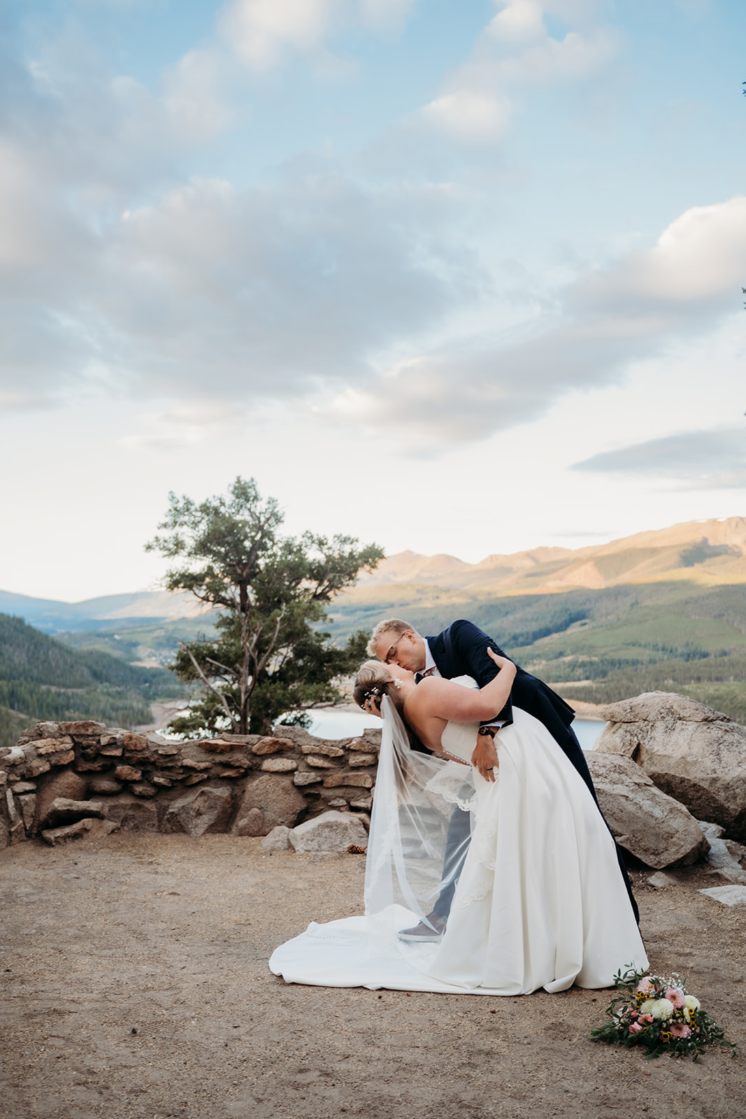 Groom dipping bride for a romantic kiss in front of mountains and lake after their ceremony.
