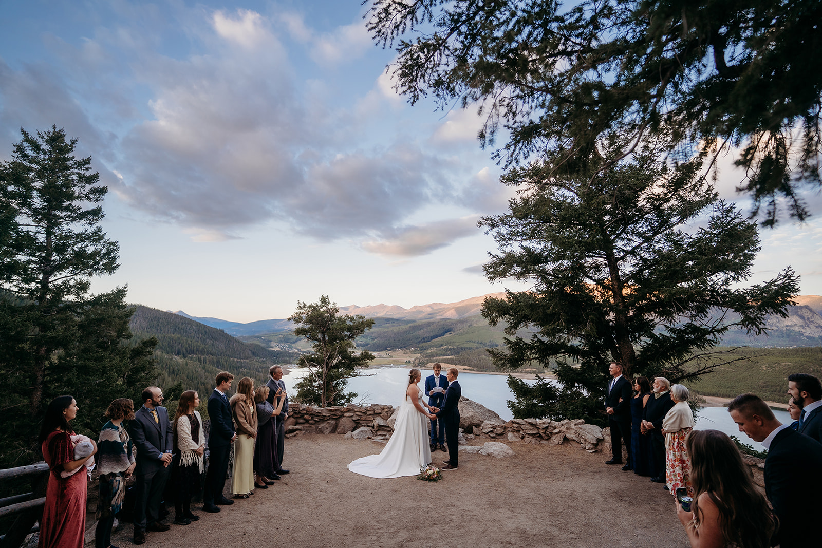 Wide mountain view of couple exchanging vows surrounded by family, lake and peaks glowing at sunset, photographed by a Denver elopement photographer.