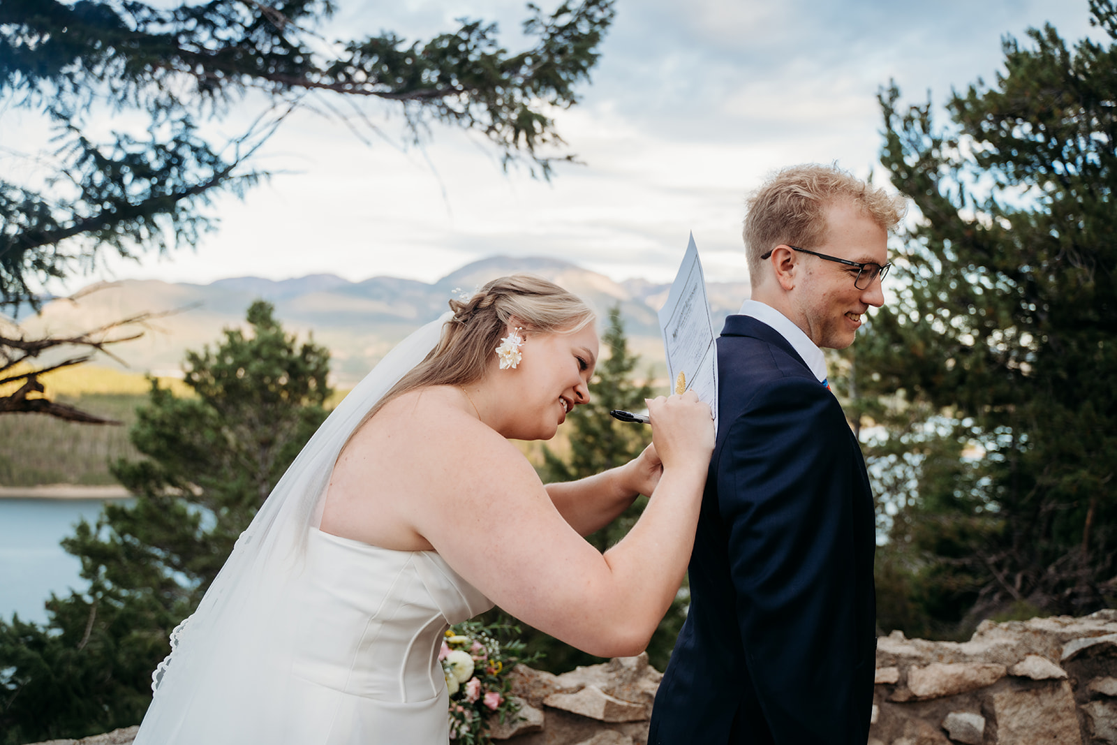 Bride jokingly writing on groom’s back with paper and pen, candid lighthearted wedding moment outdoors.