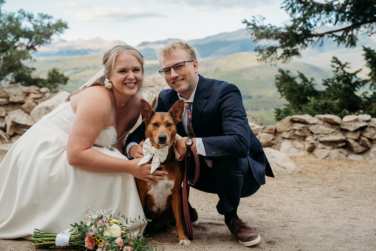 Bride and groom kneeling with their wedding dog wearing a bow, all smiling at the camera with mountain backdrop.