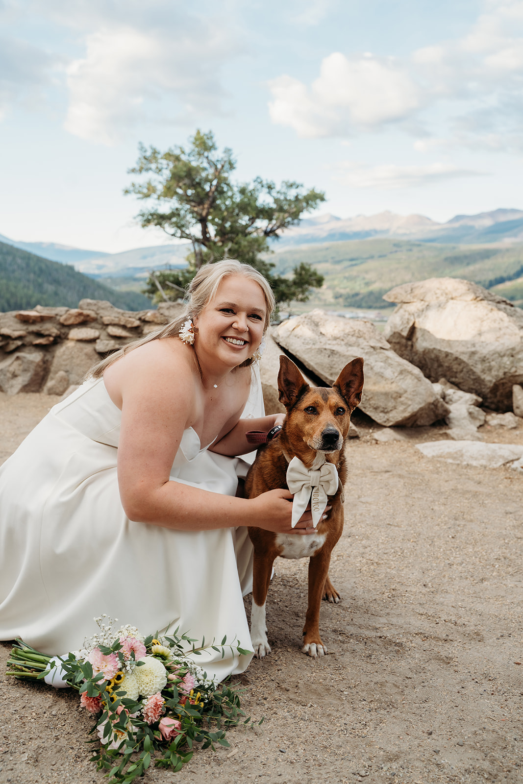 Smiling bride kneeling with her dog, both posing happily at their mountain wedding ceremony site.