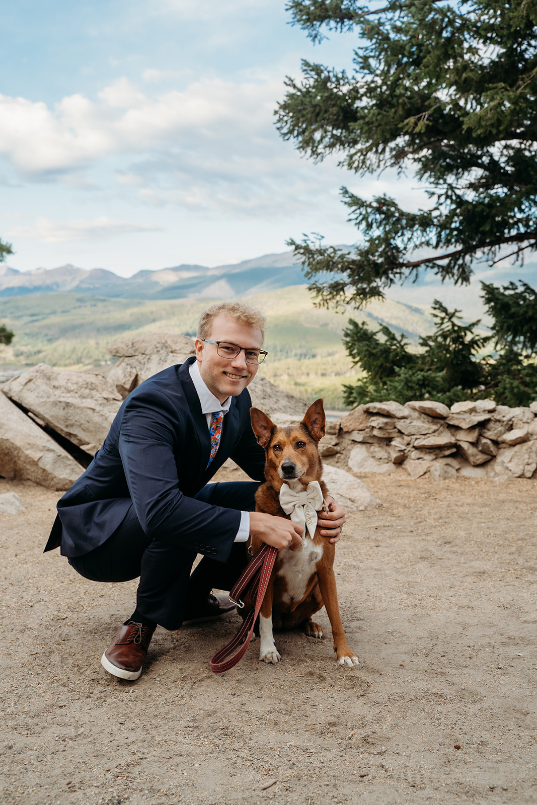 Groom kneeling next to wedding dog with mountains in the background, outdoor ceremony moment by Denver elopement photographer.