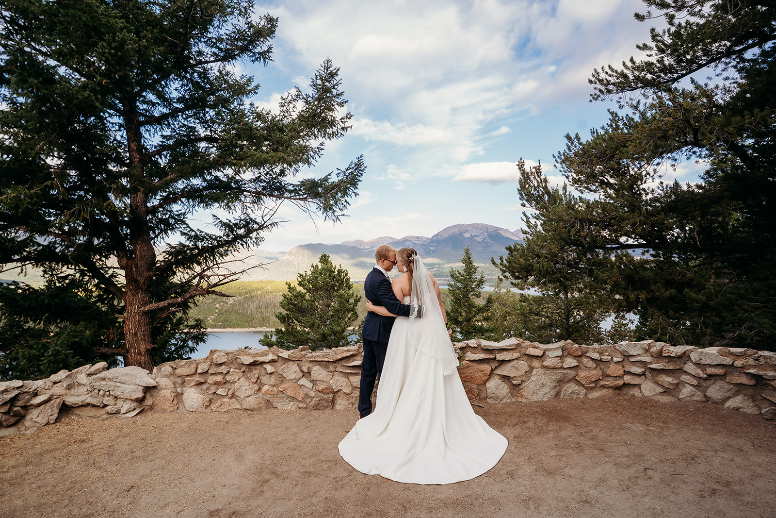 Bride and groom embracing with mountains, trees, and lake in the distance, captured from behind for a scenic wedding portrait.