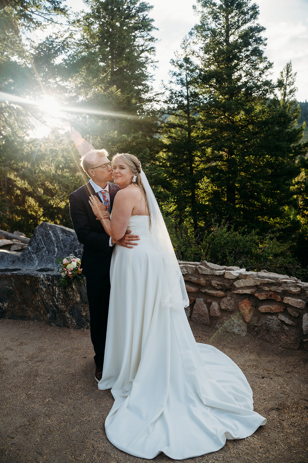 Bride and groom embracing with sunlight streaming through pine trees, captured by a Denver elopement photographer.