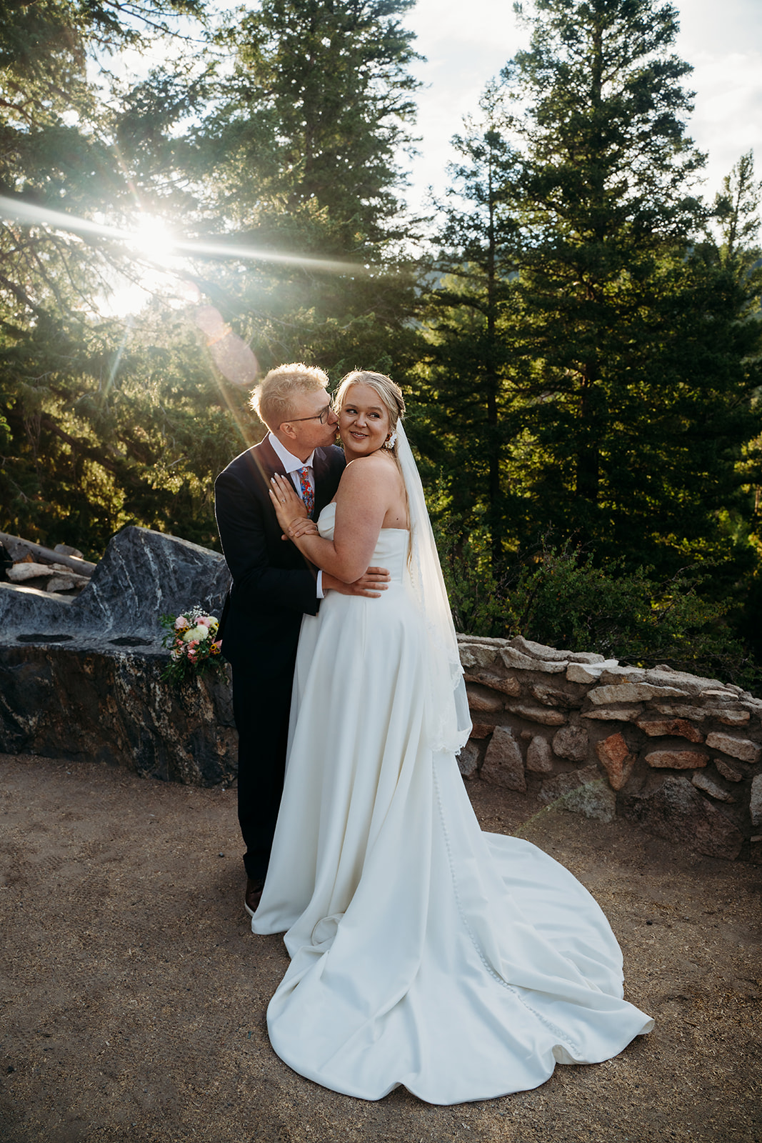 Groom kissing bride’s cheek as sunlight filters through the pine trees, joyful moment by a Denver elopement photographer.