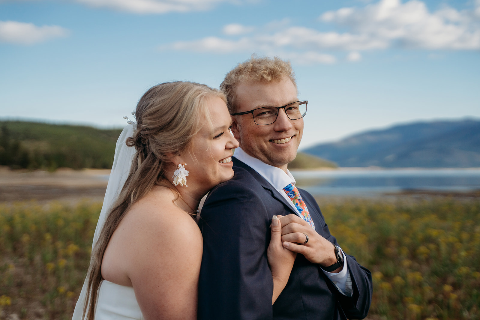 Bride and groom laughing together, holding hands with lake and mountains in the background, captured by a Denver elopement photographer.