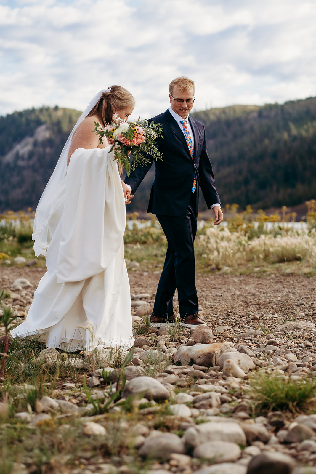 Bride and groom holding hands while walking across a rocky field, bouquet in hand, mountain backdrop, by Denver elopement photographer.