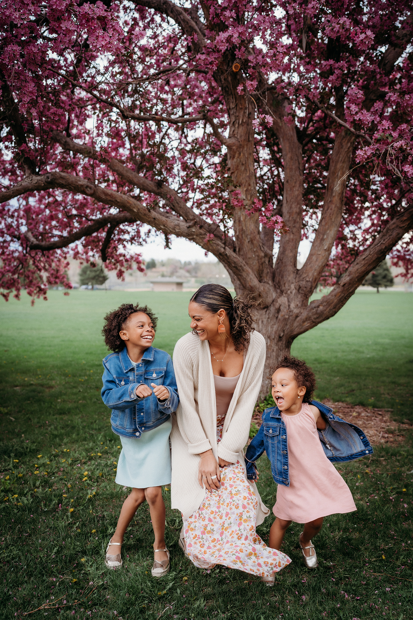 Mom with two daughters in pastel dresses and jean jackets under a flowering tree, a fun family portrait.