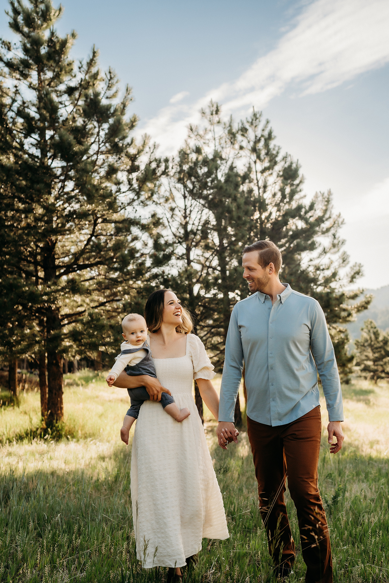 Couple walking with their baby in the mountains, dressed in light neutral outfits for family photos.
