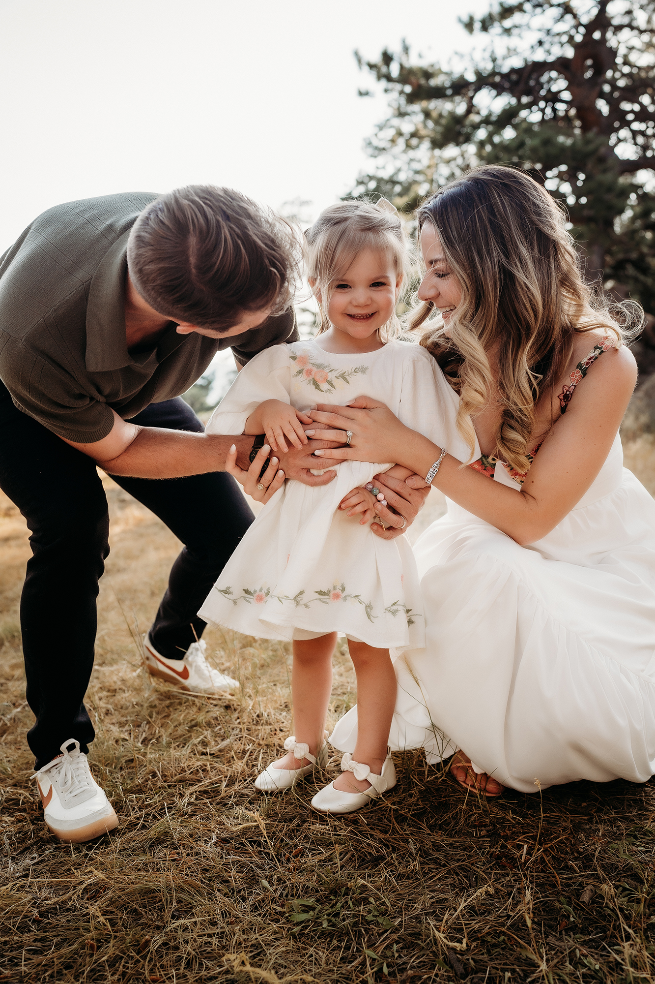 Parents crouched down with their daughter in a white floral dress, sharing a candid family moment.