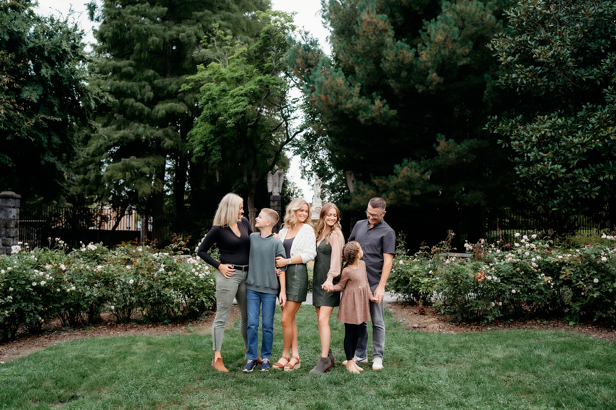 Large family standing together in a garden, dressed in shades of green, black, and neutral tones.
