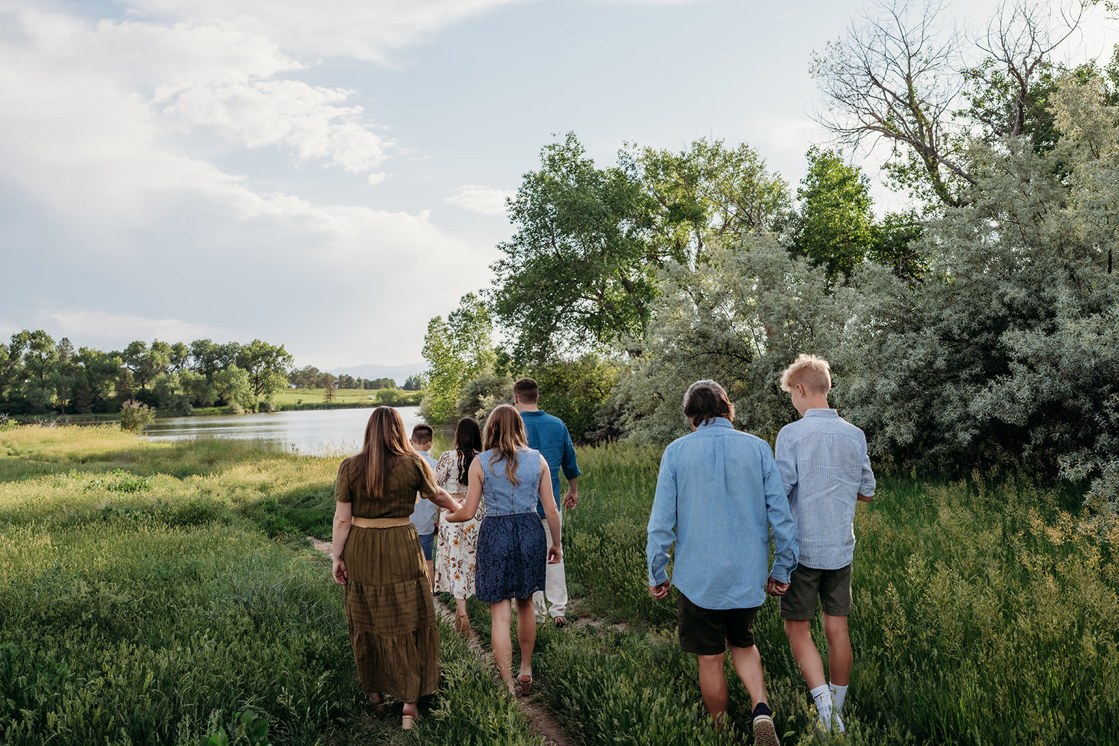 Family walking hand in hand down a path toward a lake, dressed in light summer outfits.