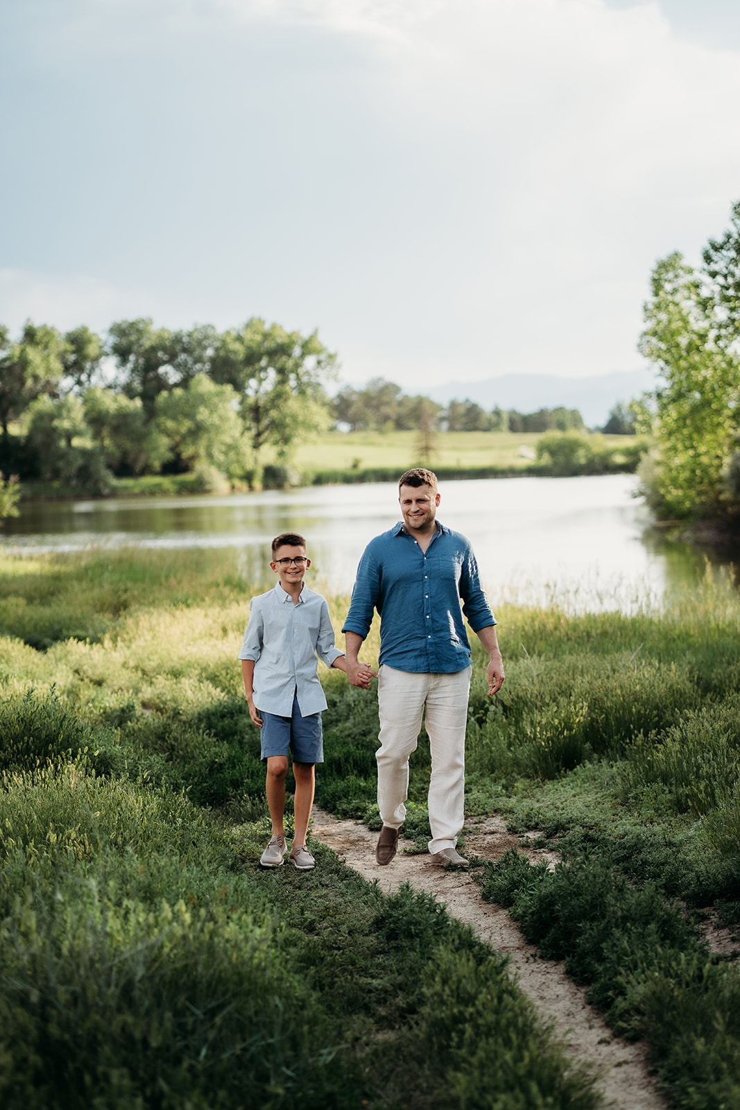 Father and son walking hand in hand on a trail, dressed in light blue shirts and neutral tones.