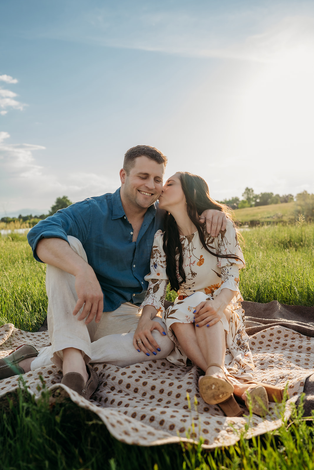 Couple sitting on a blanket in the grass, wearing romantic and relaxed outfits during sunset.