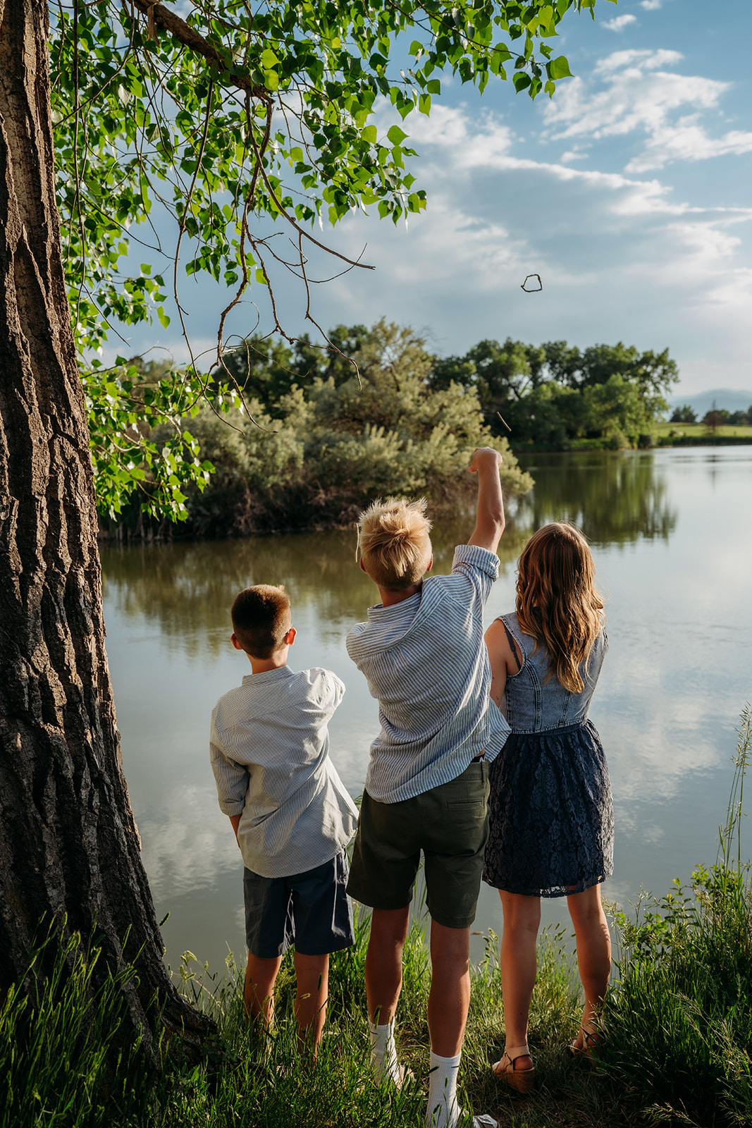 Three siblings facing a lake, throwing rocks while dressed in coordinated summer outfits.