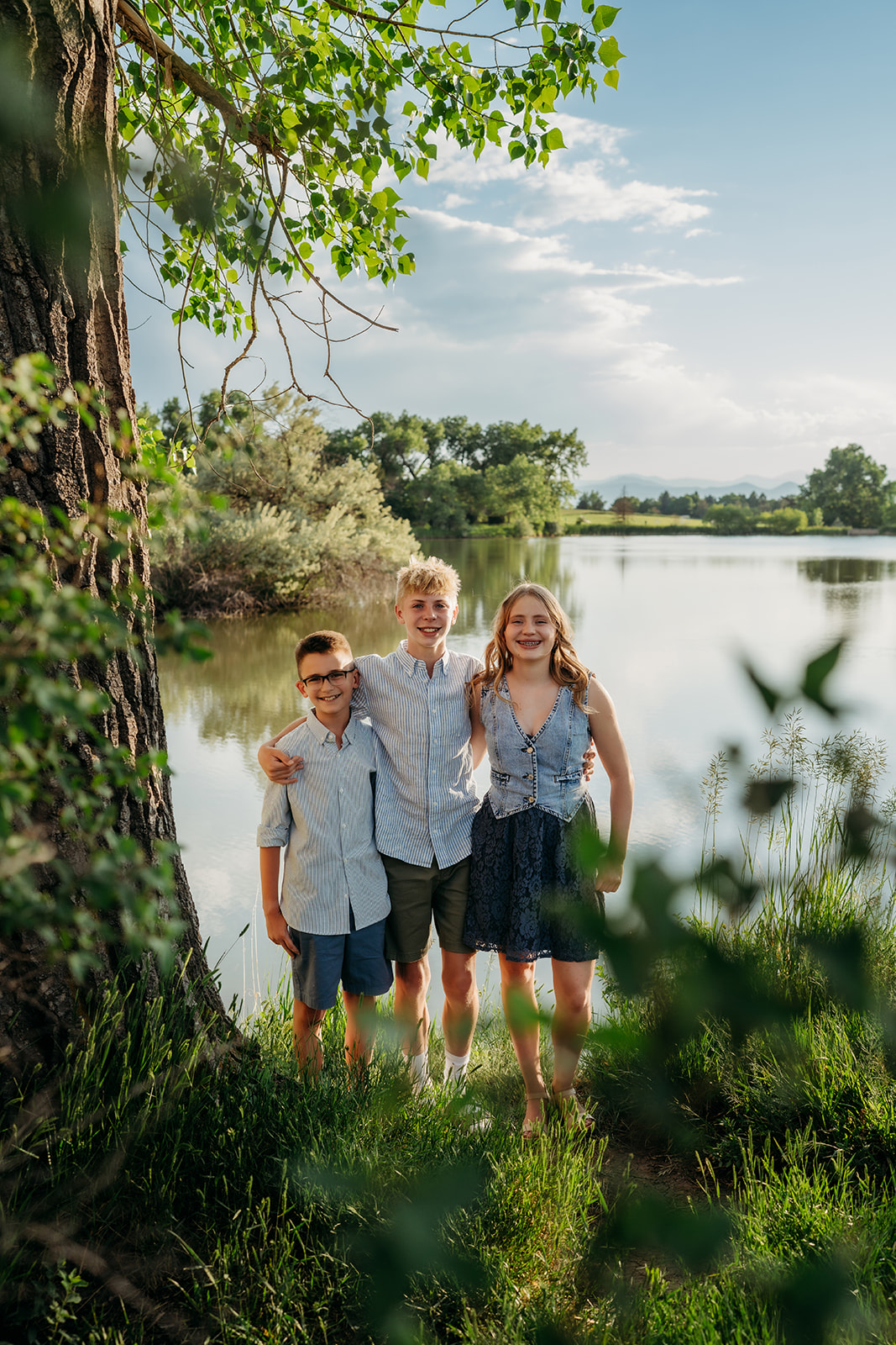 Three siblings smiling together near a lake, dressed in light and coordinated outfits for family photos.