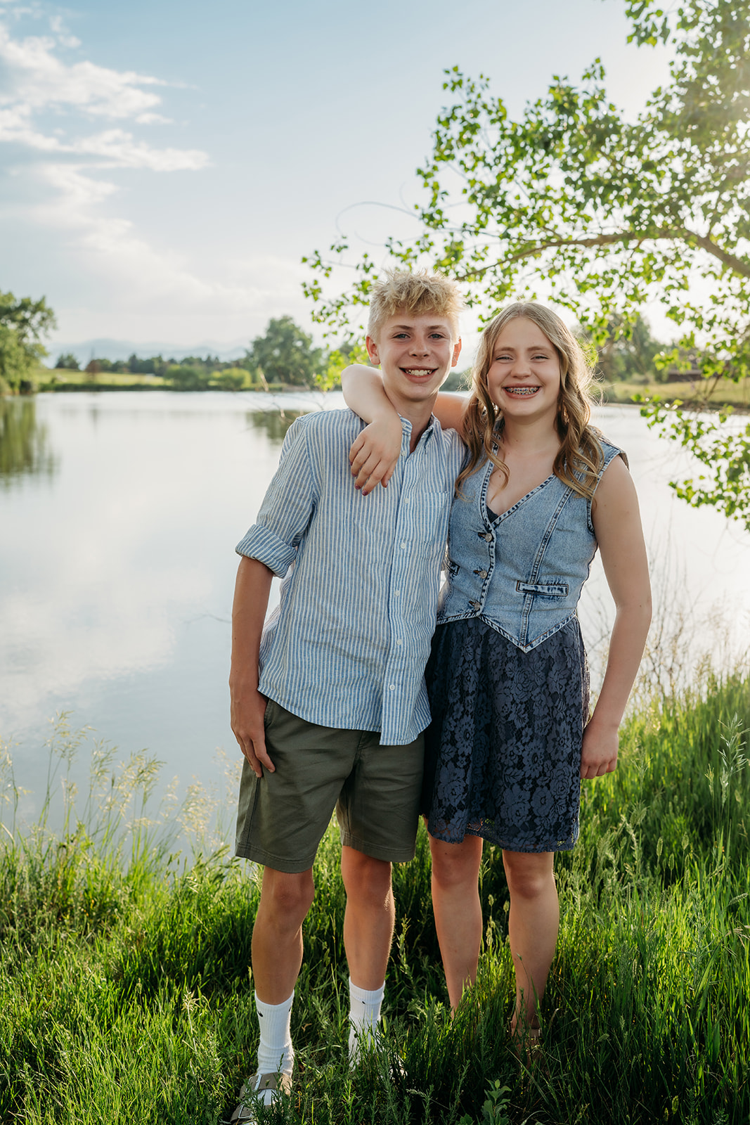 Brother and sister smiling by a lakeside, wearing casual summer outfits for family photos.