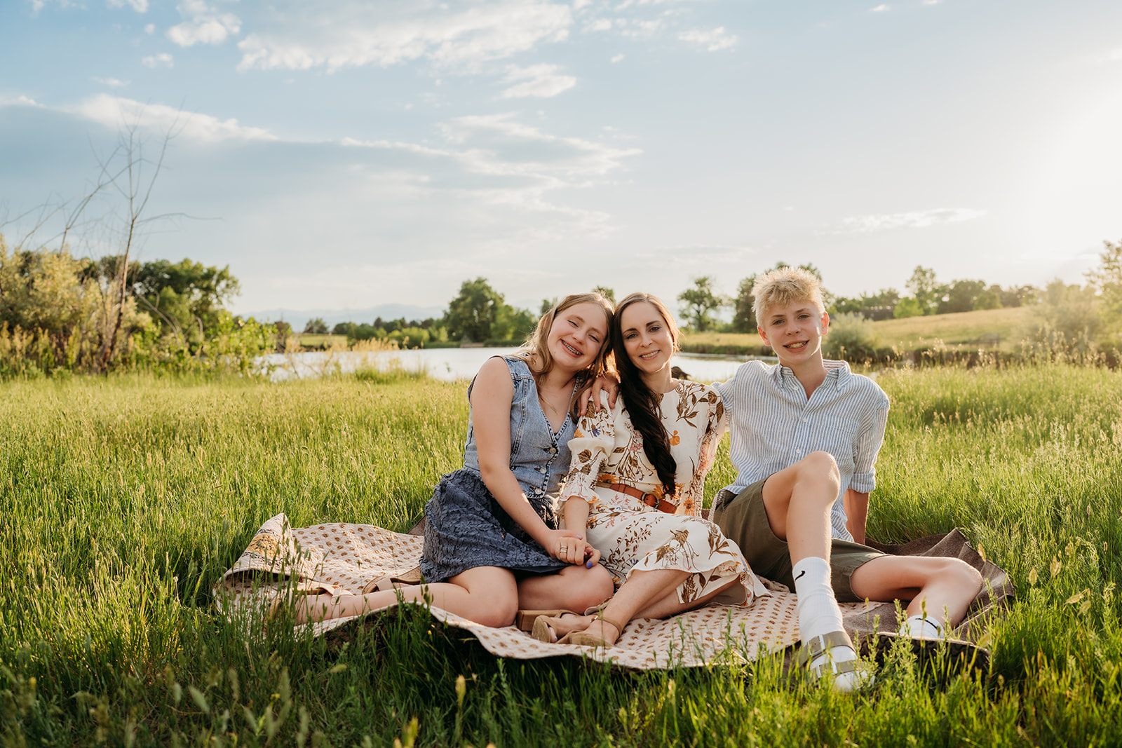Two siblings with their mom sitting on a blanket in a grassy field, showcasing soft-toned outfits for family portraits.