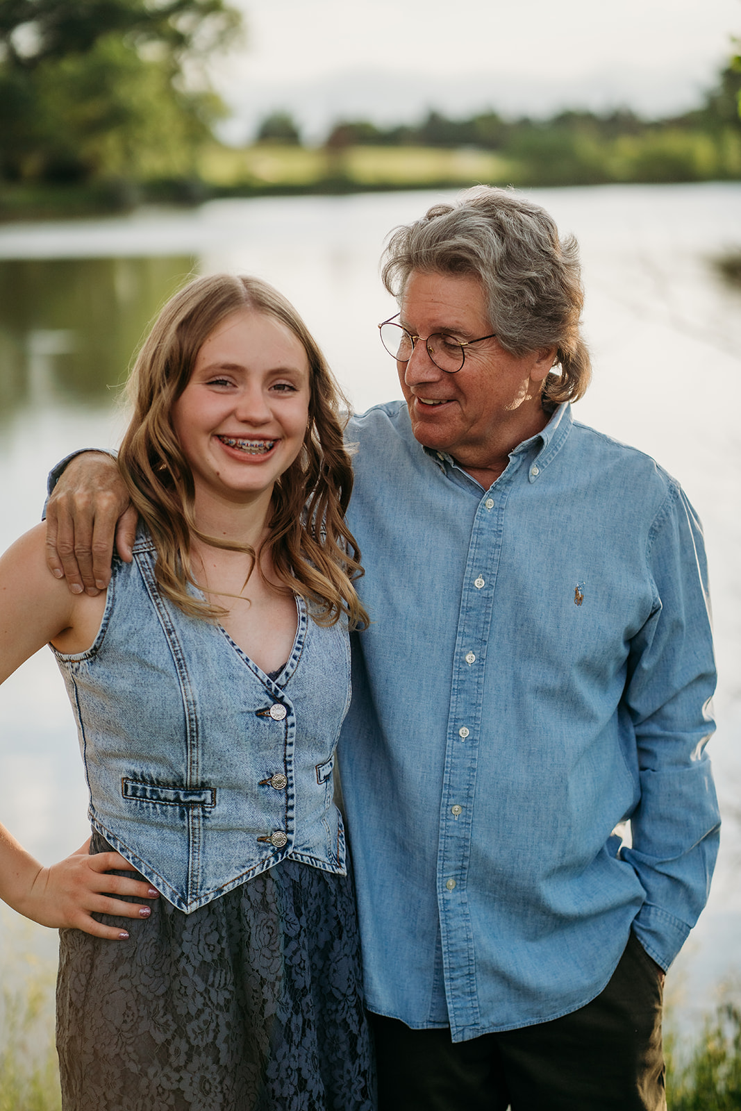 Grandfather standing with his granddaughter by the lake, both dressed in casual denim outfits.