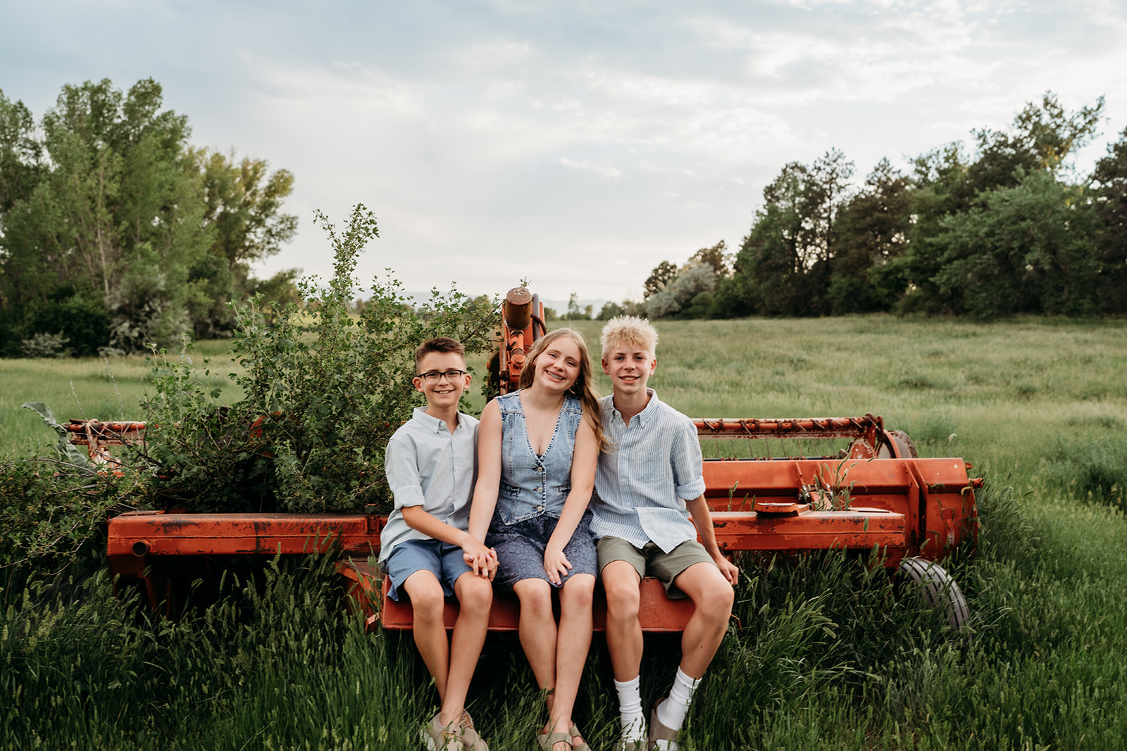Three siblings sitting on old farm equipment, dressed in coordinated blue and neutral outfits for family photos.
