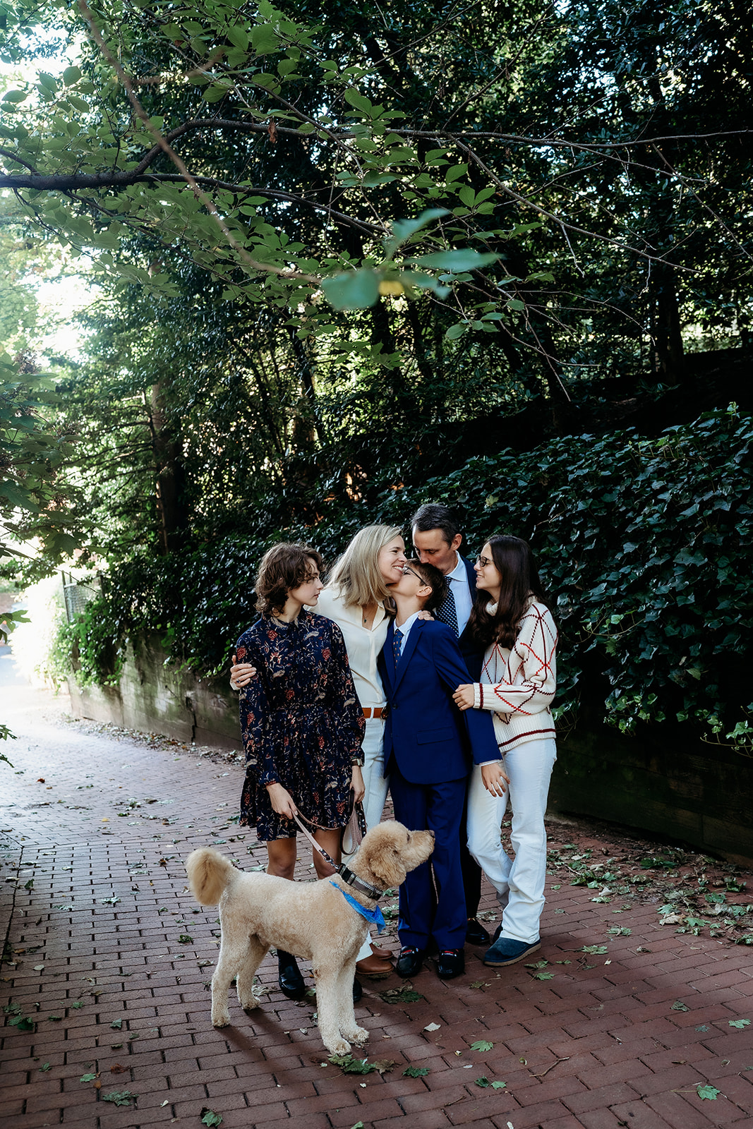 The whole family gathers under the trees with their dog, smiling and hugging in the middle of a red brick path.