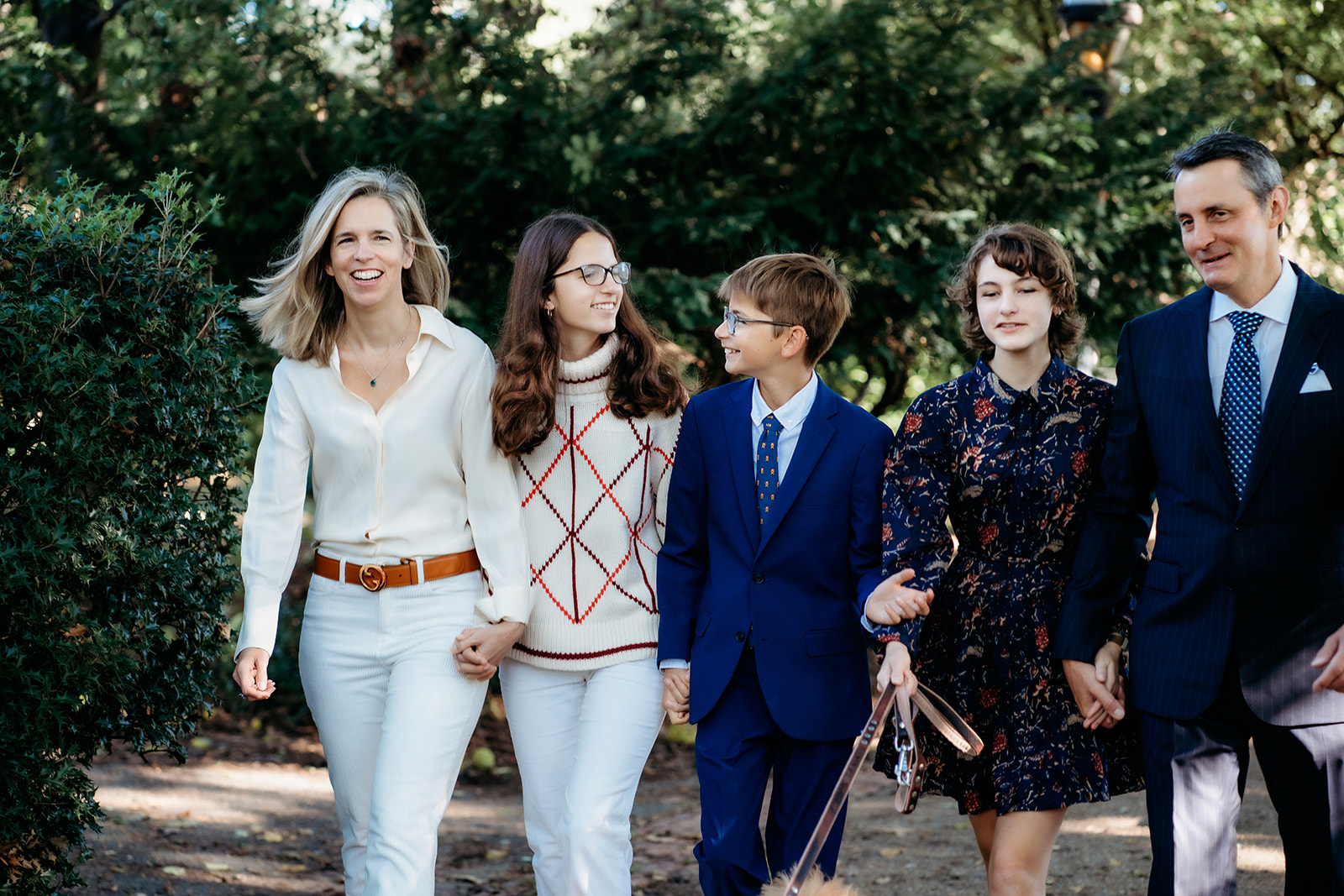 Family walks side by side through a shaded park path, laughing together during their annual family photo session.