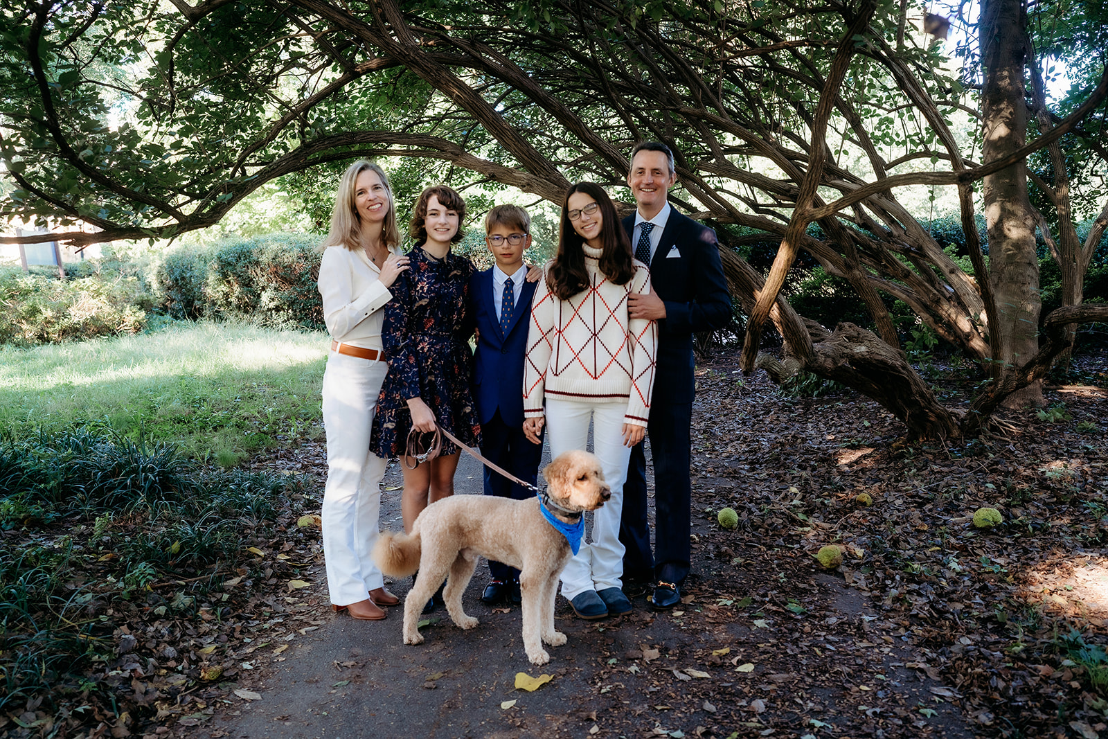 Group stands beneath a canopy of trees with their dog, dressed in coordinated fall outfits for their annual family photos.