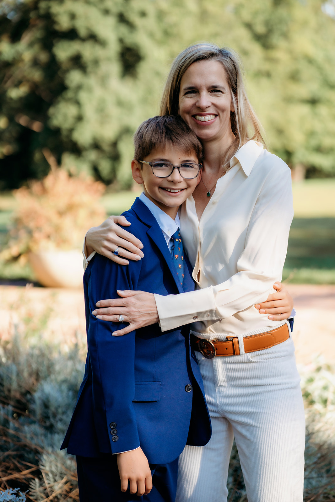 Mom wraps her arms around her son during their annual family photos, both smiling warmly in a sunlit garden setting.