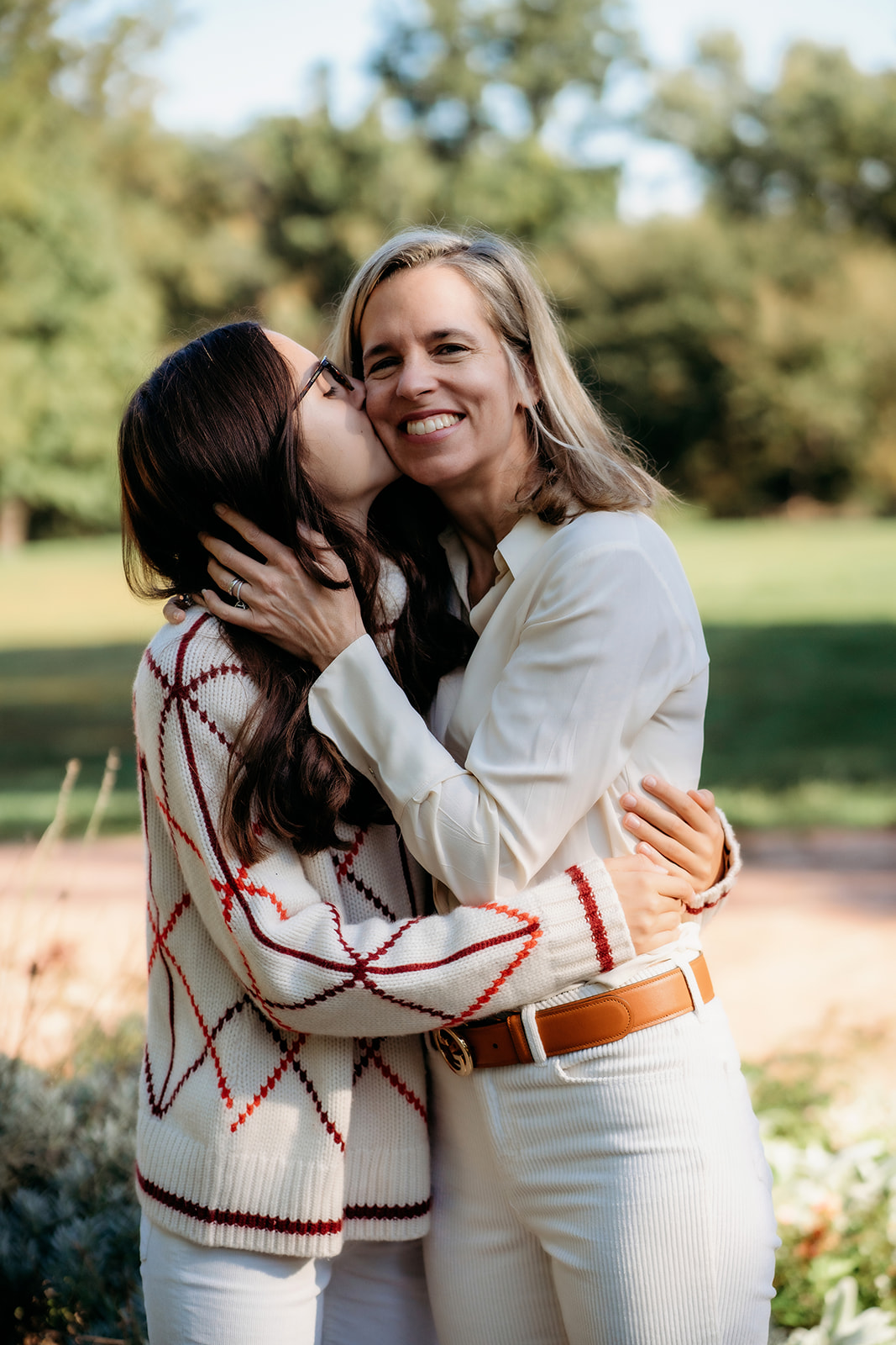 Daughter kisses her mom on the cheek during their annual family photos, sharing a sweet moment outdoors in soft morning light.