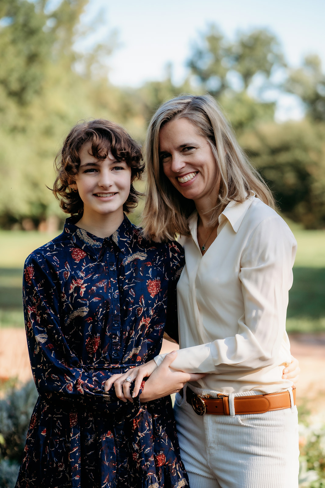 Mom and daughter hold hands and smile softly, standing together in a sunny garden scene filled with early autumn color.