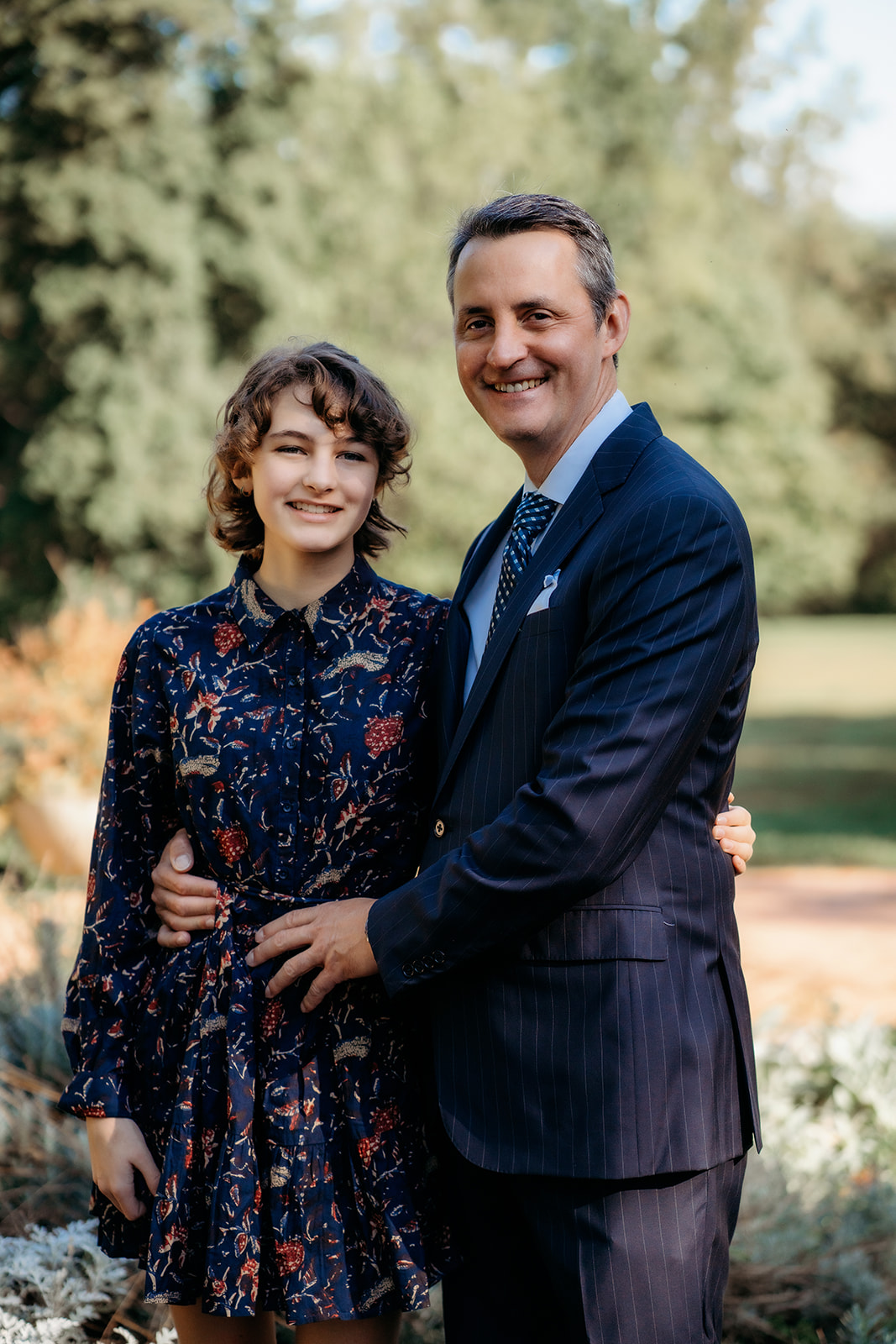 Dad wraps his arm around his daughter in a floral dress, standing together in front of soft green trees during their family shoot.