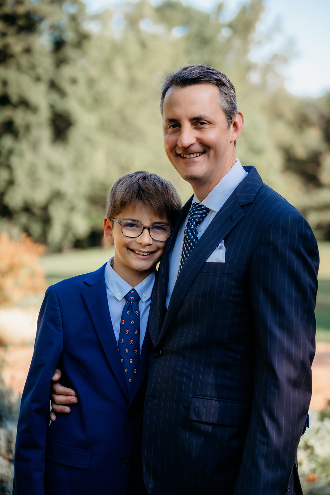 Father and son dressed in matching navy suits smile and hug during a special moment from their annual family photo session.
